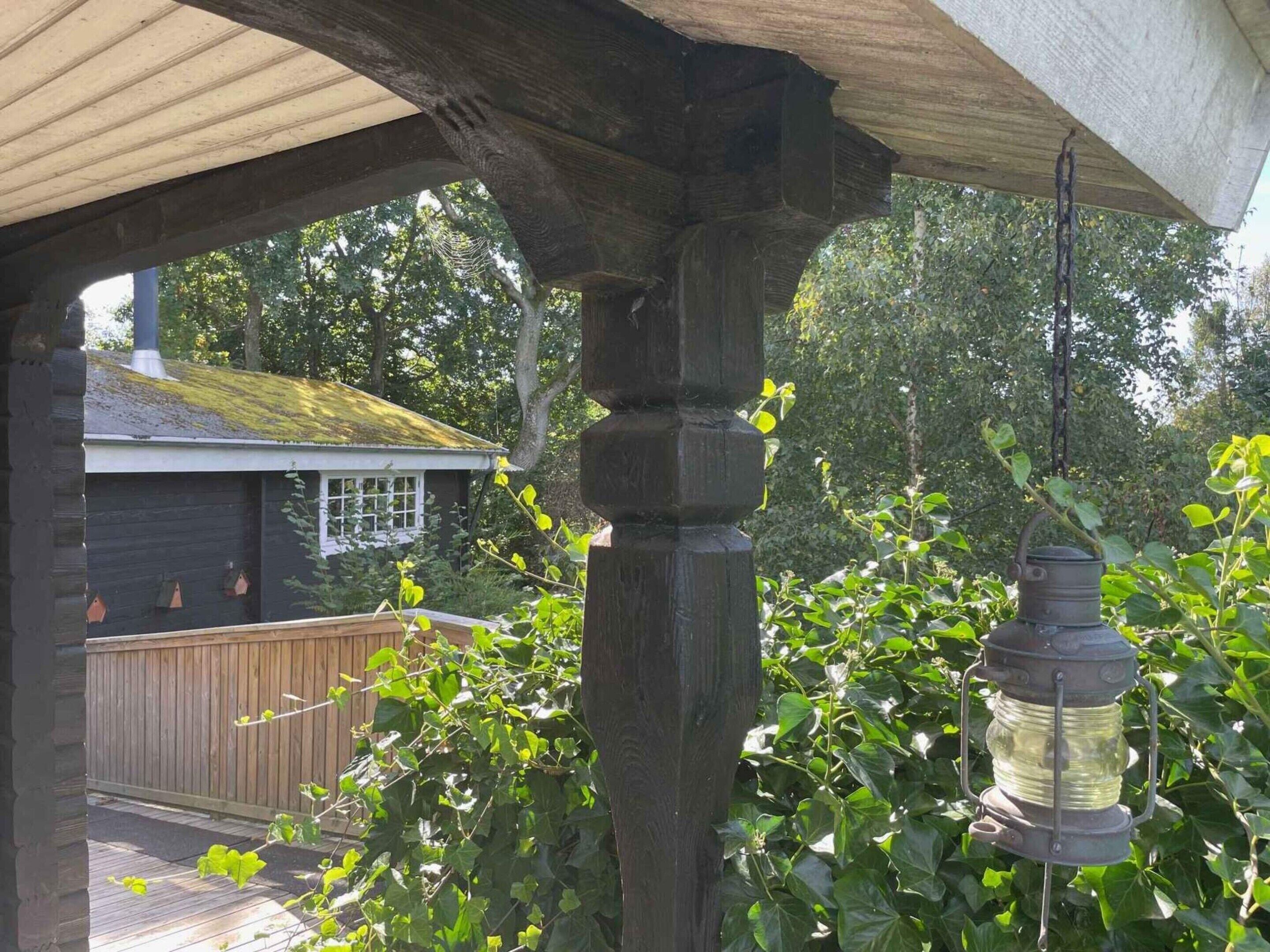 A wooden pergola structure with a hanging lantern, surrounded by lush greenery and a wooden deck.