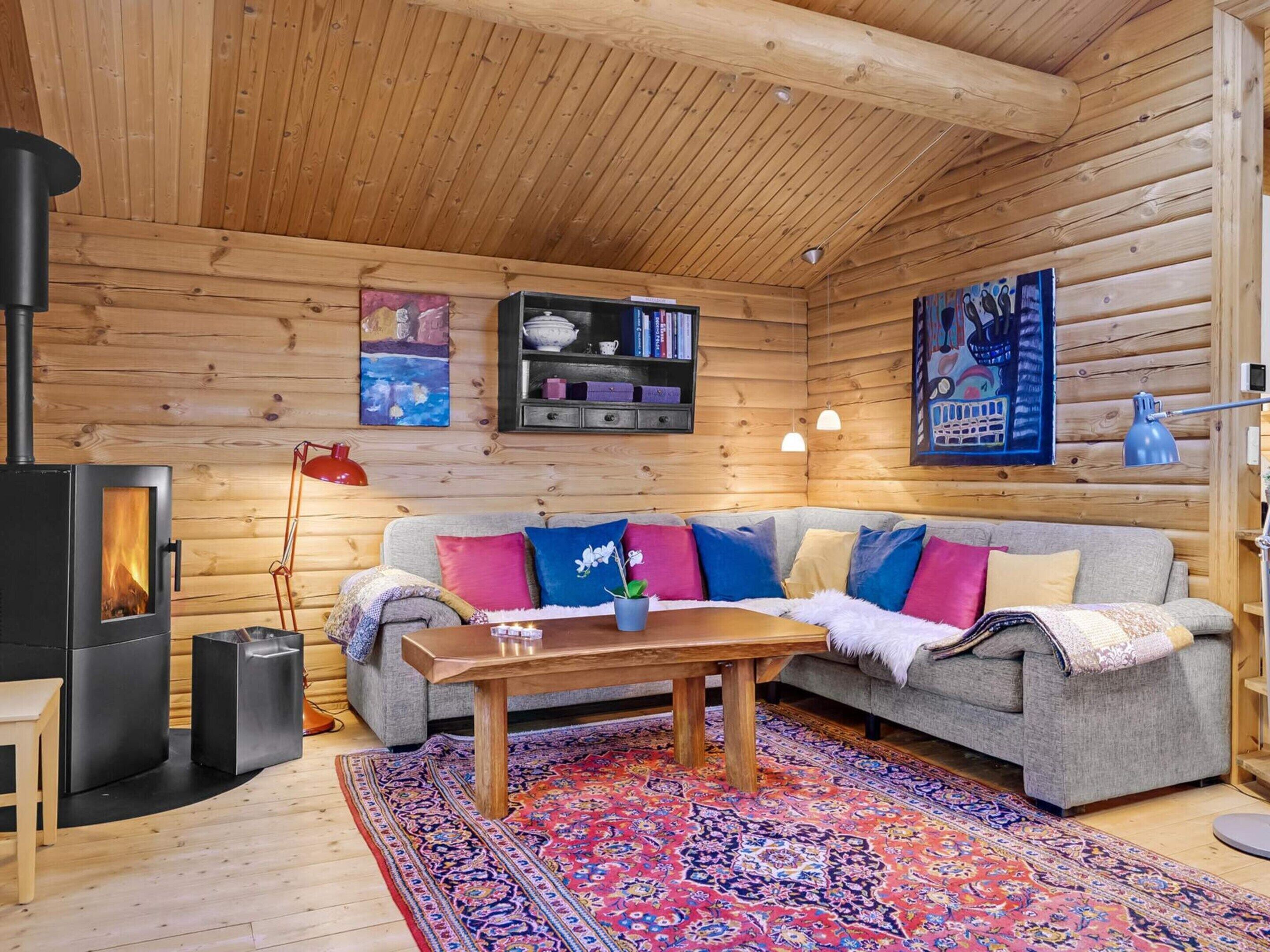 A cozy log cabin interior with a fireplace, a sofa with colorful cushions, a wooden coffee table, and a patterned rug.