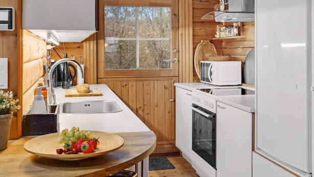 A compact kitchen with wooden walls, a window, a sink, a microwave, and a refrigerator. - 5 Star Holiday Home in Glesborg (Glesborg)