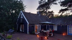 En træterrasse med udendørs møbler, en overdækket terrasse og et hus med mørkt tag og hvide detaljer.