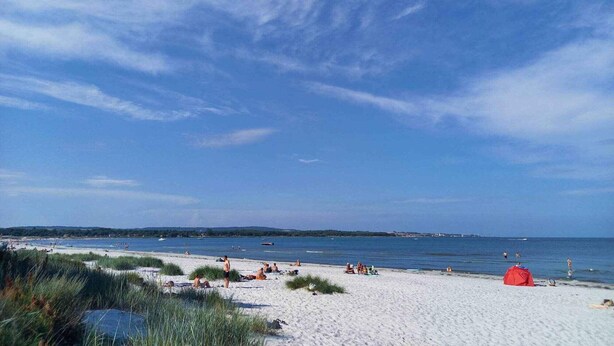 En strand med folk, der solbader og svømmer, et rødt telt og en klar blå himmel.