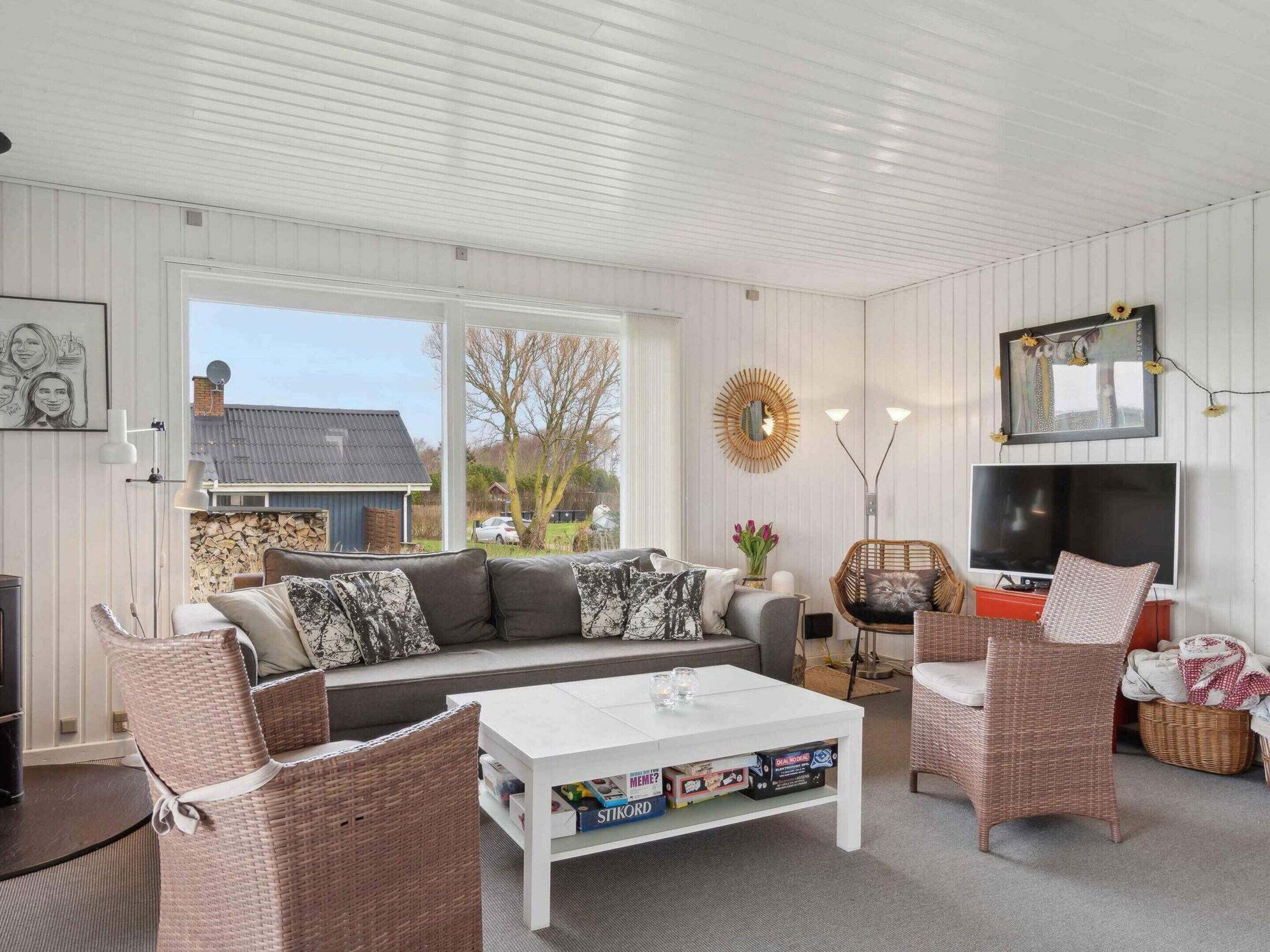 Ein gemütliches Wohnzimmer mit einer Couch, zwei Korbstühlen, einem Couchtisch mit Gesellschaftsspielen, einem Fernseher und einem großen Fenster mit Blick auf ein Haus und Bäume.