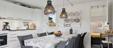 A modern kitchen with white cabinets, a dining table with a patterned tablecloth, wicker chairs, and pendant lights.