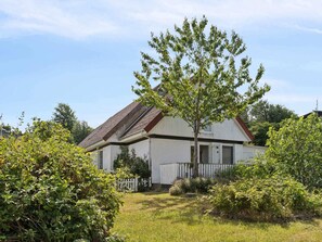 A single-story house with a sloped roof, surrounded by a white picket fence and various shrubs.