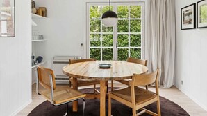 En matplats med ett runt träbord, fyra trästolar, en hängande taklampa och ett stort fönster med utsikt över grönskan.