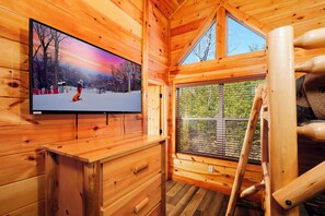 A wooden cabin interior with a large TV on a wooden cabinet, a window with a view of trees, and a wooden ladder.