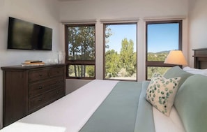 A bedroom with a bed, a dresser, a TV, and large windows with a view of trees and hills.