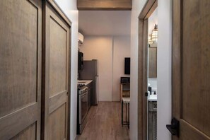 A narrow hallway with wooden doors, a kitchen area with a stove and refrigerator, and a small bar stool.