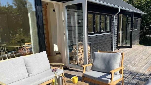 A wooden deck with a sofa and chairs, a table with a vase and mug, and a glass door leading to a room with a fireplace. - 5 Star Holiday Home in Vejby (Vejby)