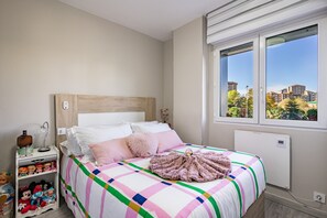 A bedroom with a bed, a nightstand, a window, and a view of buildings outside.