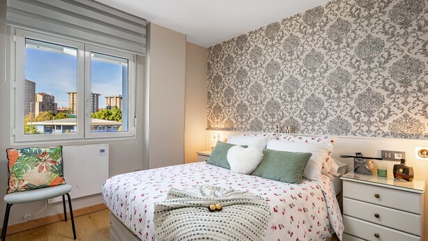 A bedroom with a bed, a chair, a nightstand, and a window with a view of buildings.