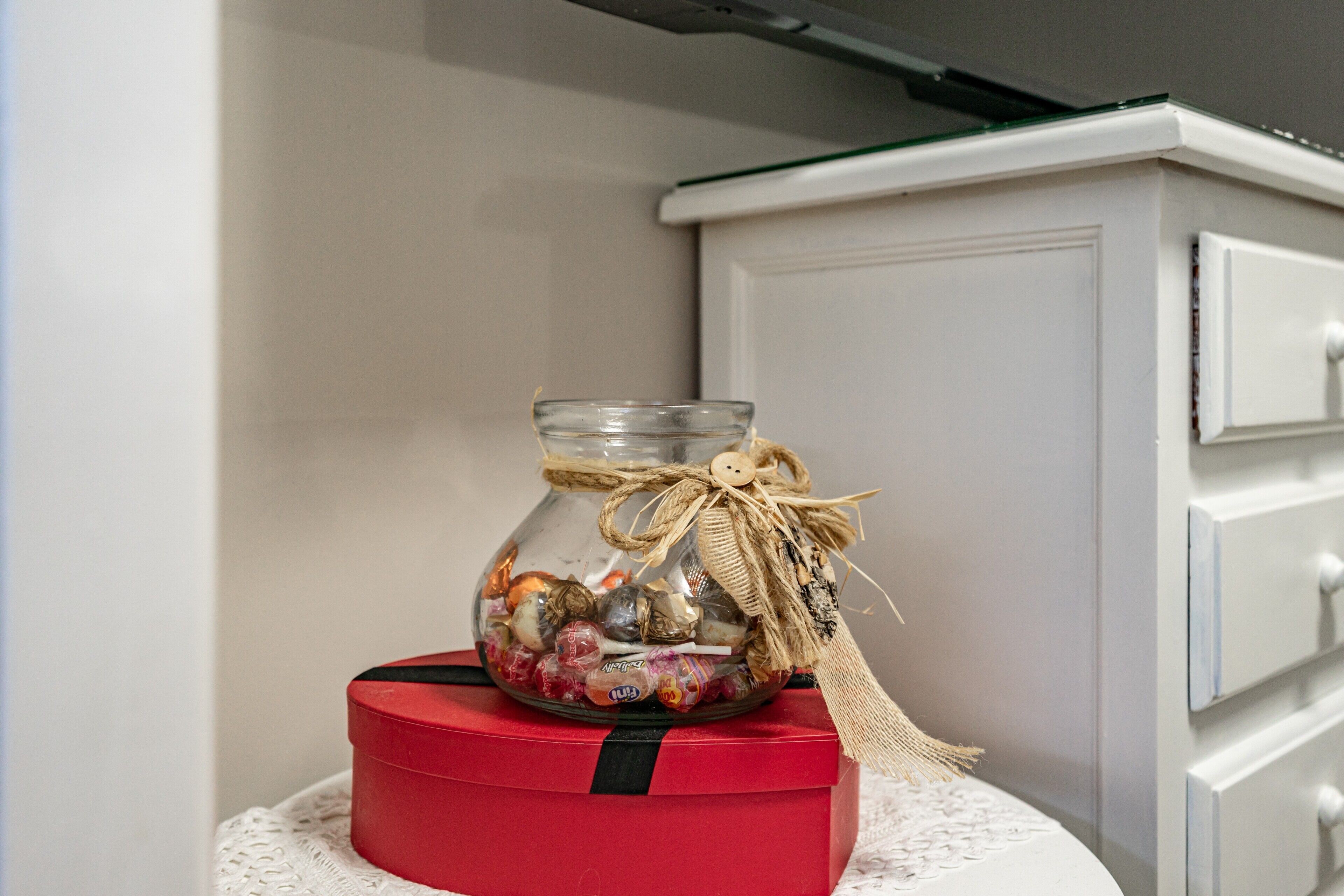 A clear glass jar with a burlap ribbon and a button, filled with assorted candies, placed on a red box with a black stripe, against a white wall and a white cabinet.
