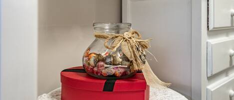 A clear glass jar with a burlap ribbon and a button, filled with assorted candies, placed on a red box with a black stripe, against a white wall and a white cabinet.