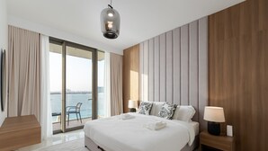 Een moderne hotelkamer met een groot bed, een nachtkastje met lamp, uitzicht op zee door middel van kamerhoge ramen en een balkon met een stoel.