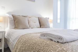 Un lit bien fait, avec une couette blanche et une couverture enroulée, une lampe de chevet et une fenêtre avec des rideaux.