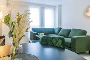 Ein modernes Wohnzimmer mit einem grünen Sofa, einer Glaskaraffe mit Trockenpflanzen auf einem Tisch und einem Fenster mit Tageslicht.