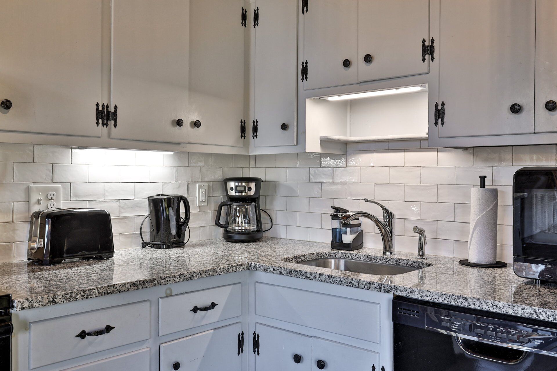 A modern kitchen with white cabinets, a granite countertop, a coffee maker, a toaster, a sink, and a dishwasher.