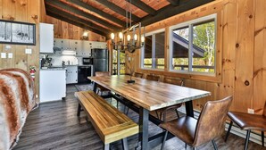 A cabin interior with a wooden dining table, benches, a kitchen area, and large windows offering a view of the outdoors. - RoundTop Chalet | Hot Tub, Fire Pit & Mountain Views near Killington & Okemo (Plymouth)