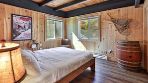 A wooden cabin room with a bed, two windows, a poster, and a rustic barrel. - RoundTop Chalet | Hot Tub, Fire Pit & Mountain Views near Killington & Okemo (Plymouth)