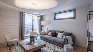 Una sala de estar moderna con piso de madera, un sofá gris, un sillón blanco, una mesa de centro de madera y un ventanal con vistas a montañas.