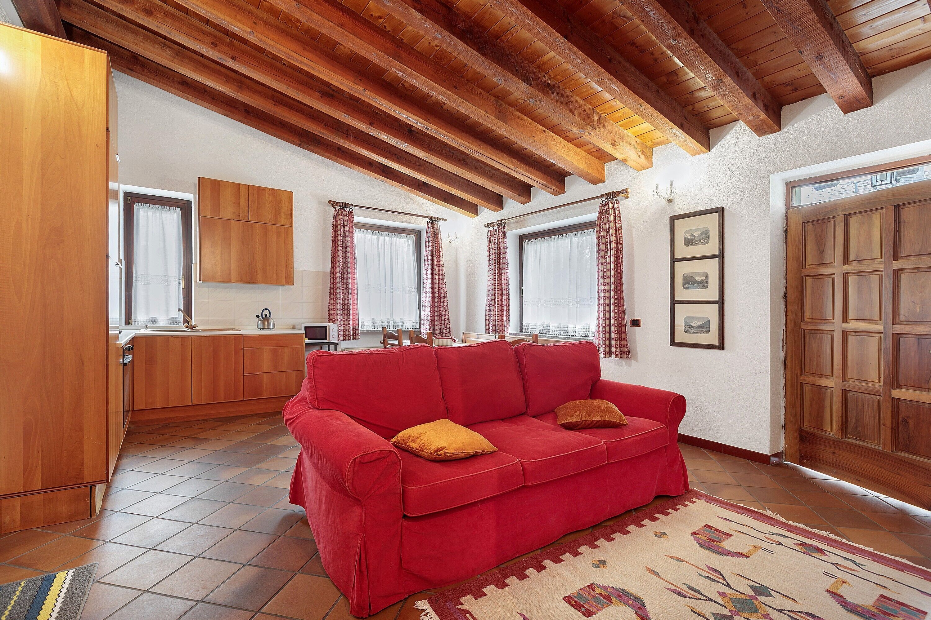 A red sofa with cushions, wooden ceiling beams, a kitchen area, and a patterned rug.