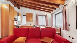 Un sofá rojo con cojines, armarios de madera, una zona de cocina y un ventanal con cortinas.
