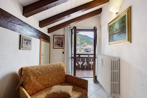 Une pièce avec un plafond en bois, un radiateur, un canapé et une porte donnant sur un balcon d’où l’on peut admirer des maisons et des montagnes.