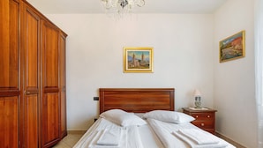 Een slaapkamer met een houten garderobe, een bed met witte lakens, een nachtkastje met een lamp, en twee ingelijste foto’s aan de muur.
