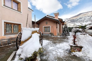 A snow-covered courtyard with a wooden cart, a building with a chimney, and a mountain in the background.