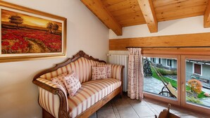 Techo de madera, un sofá con cojines estampados, un cuadro enmarcado y vistas a un jardín a través de una ventana.