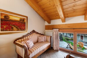 A wooden ceiling, a sofa with patterned cushions, a framed painting, and a view of a garden through a window.