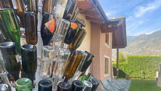Varias botellas de vino dispuestas al aire libre, con un edificio y un paisaje montañoso al fondo.