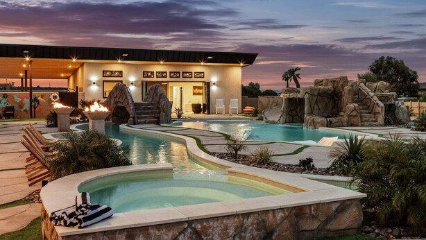 A pool area with a waterfall feature, a covered seating area, and a building with a modern design. - The Lazy River House - Joshua Tree Resort (Joshua Tree)