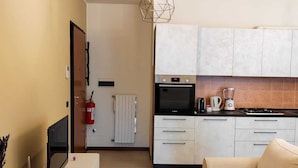 Une cuisine moderne avec des meubles blancs, un four noir et une machine à café. Le salon est équipé d’un canapé beige et d’un téléviseur.