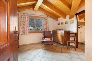 Una habitación con techo de madera, una ventana con cortinas, una silla, un aparador y un cuadro en la pared.