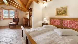 Ein Schlafzimmer mit Holzbalkendecke, einem Bett mit weißen Leinentüchern, einem Sessel, einem Schreibtisch und einem Bild an der Wand.