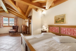 A bedroom with a wooden ceiling, a bed with white linens, a chair, a desk, and a painting on the wall.