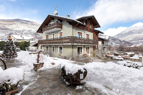 A multi-story building with balconies, a snow-covered ground, and mountains in the background.