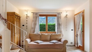 Ein Zimmer mit einer Holztreppe, einem beigefarbenen Sofa mit Kissen, einem Fenster mit Vorhängen und Blick ins Grüne.