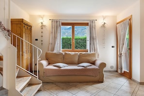 A room with a wooden staircase, a beige sofa with cushions, a window with curtains, and a view of greenery outside.