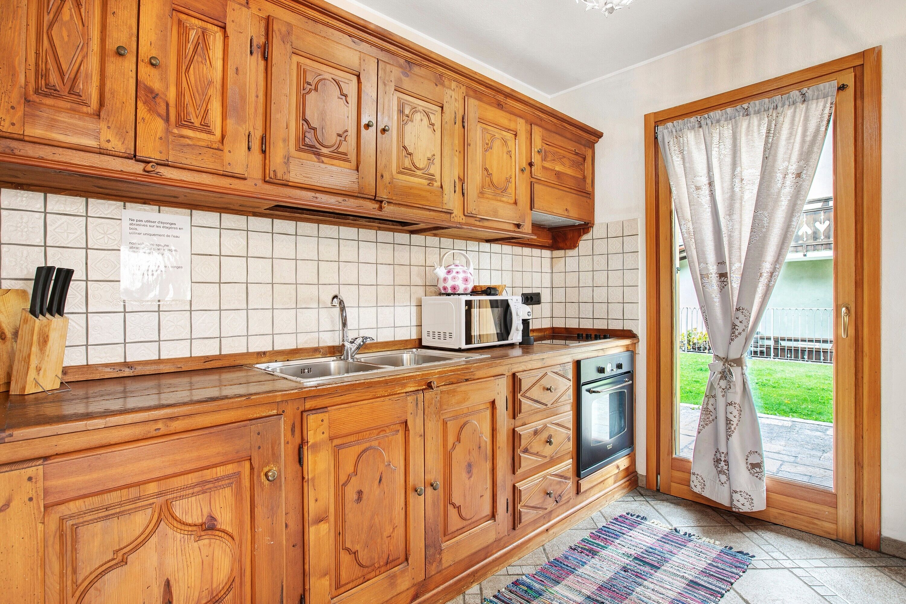 A kitchen with wooden cabinets, a microwave, a sink, and a window with a curtain.