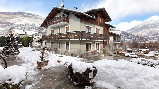 Un edificio de varios pisos con balcones, suelo cubierto de nieve y montañas al fondo.