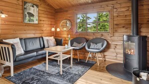 A cozy cabin interior with a wooden interior, a sofa, two armchairs, a table, a fireplace, and a window with a view of trees. - 4 Star Holiday Home in Glesborg (Glesborg)