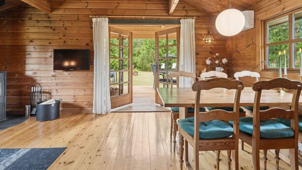 A wooden cabin interior with a dining area, a TV mounted on the wall, and a view of the outdoors. - 4 Star Holiday Home in Glesborg (Glesborg)