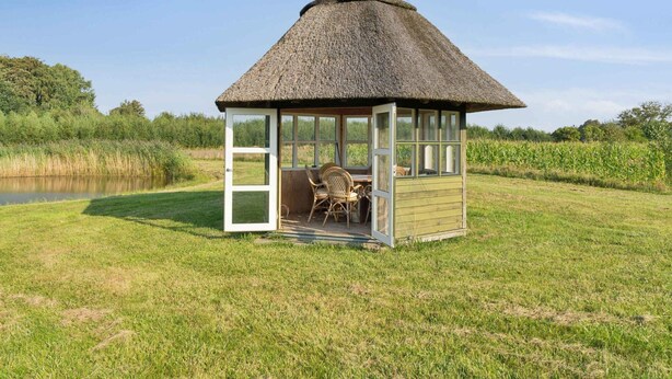 Une structure à toit de chaume dotée d’une porte et de fenêtres en verre, d’un petit espace extérieur aménagé avec des chaises, et offrant une vue sur une étendue d’eau et une végétation luxuriante en arrière-plan.
