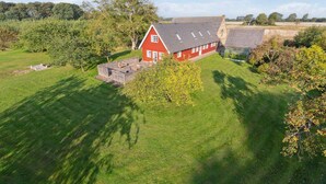 Une maison rouge avec un toit en pente et une terrasse entourée d’une clôture, le tout bordé de pelouse verte et d’arbres.