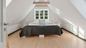 Une chambre avec un plafond en pente, un lit avec une literie blanche, une fenêtre donnant sur la verdure et une structure en poutres apparentes.