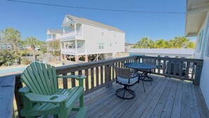 Una terraza de madera con una mesa y dos sillas Adirondack verdes, con vistas a una piscina y una casa blanca con balcón.