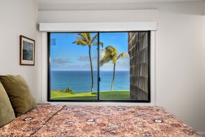 A bedroom with a large sliding glass door leading to a view of the ocean and palm trees. - Remodeled Ground Floor Ocean Views at Sealodge J5 (Princeville)