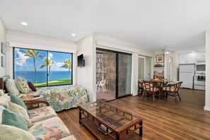 A living room with a large window overlooking the ocean, a dining area with wicker chairs, and a kitchen with white cabinets. - Remodeled Ground Floor Ocean Views at Sealodge J5 (Princeville)